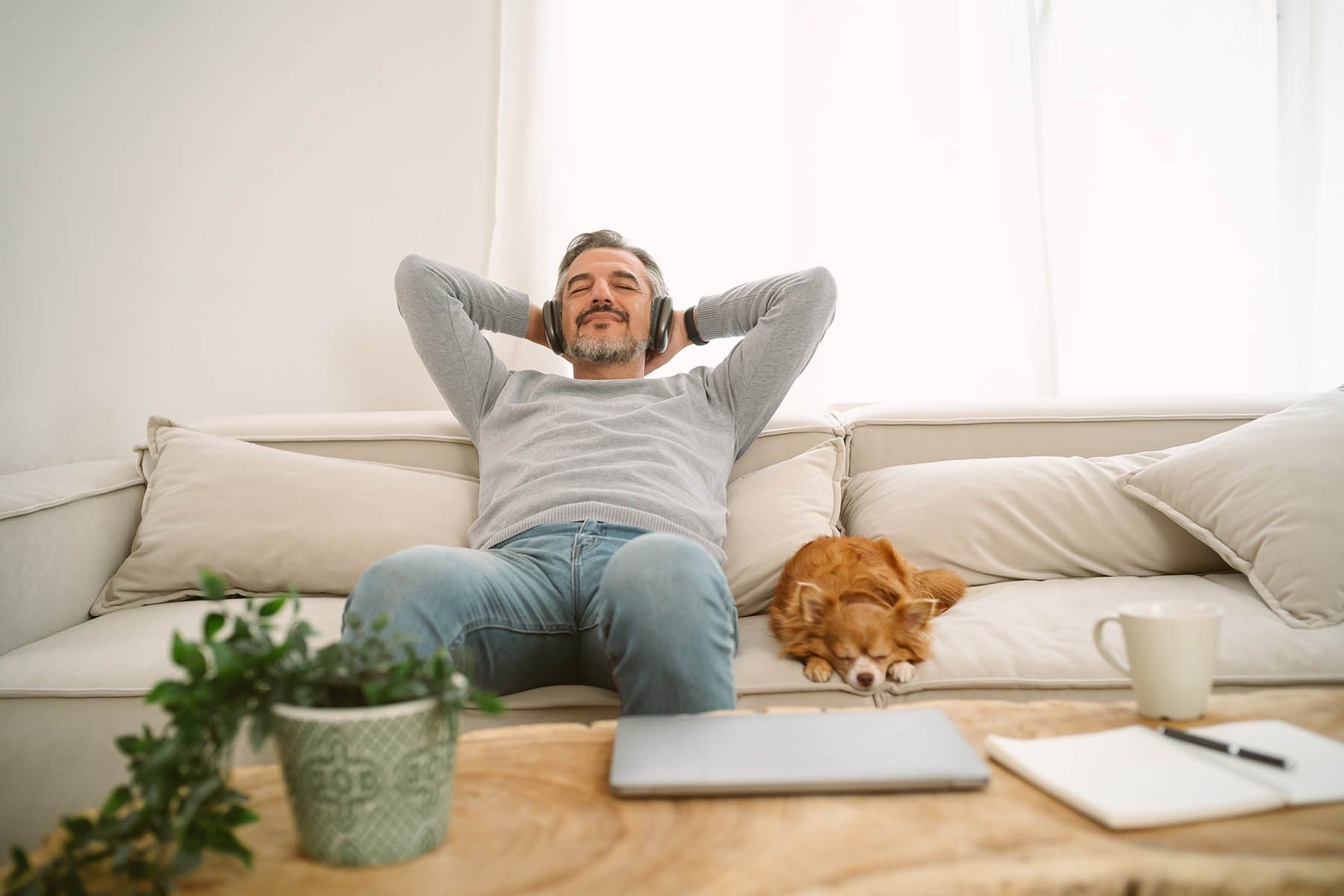 Calm Middle age Caucasian man sitting on sofa listening to music enjoying meditation for sleep and peaceful mind in wireless headphones, leaning back with his lovely chihuahua dog sit besides.